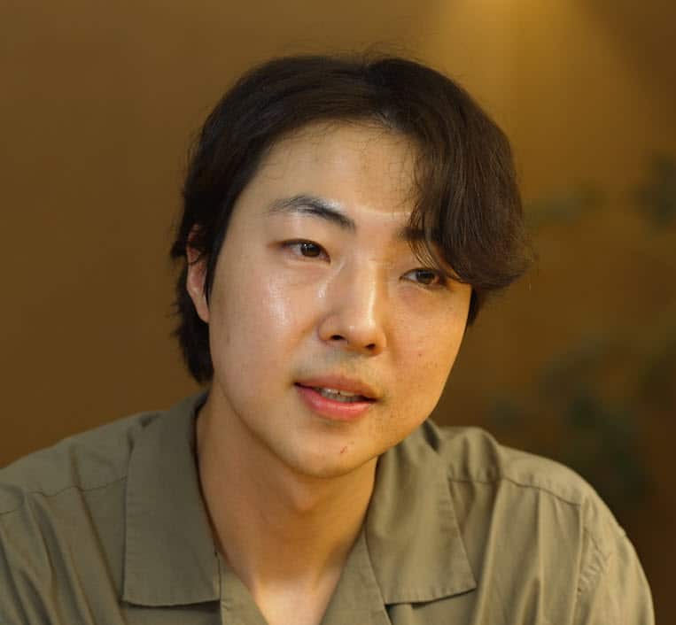 A man with dark brown hair and wearing a green shirt sits facing camera.  