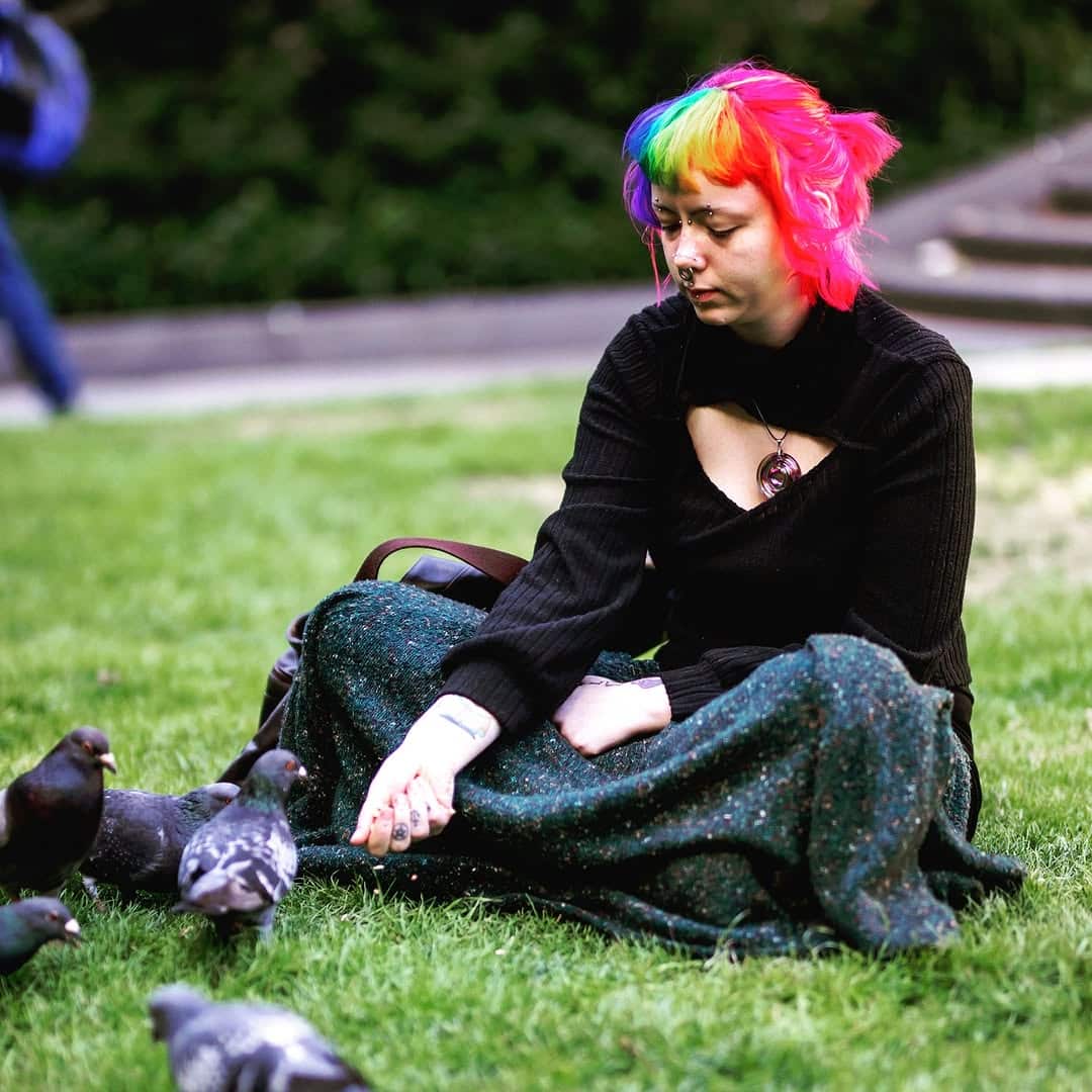 A woman with bright coloured hair sitting on the ground with pigeons.