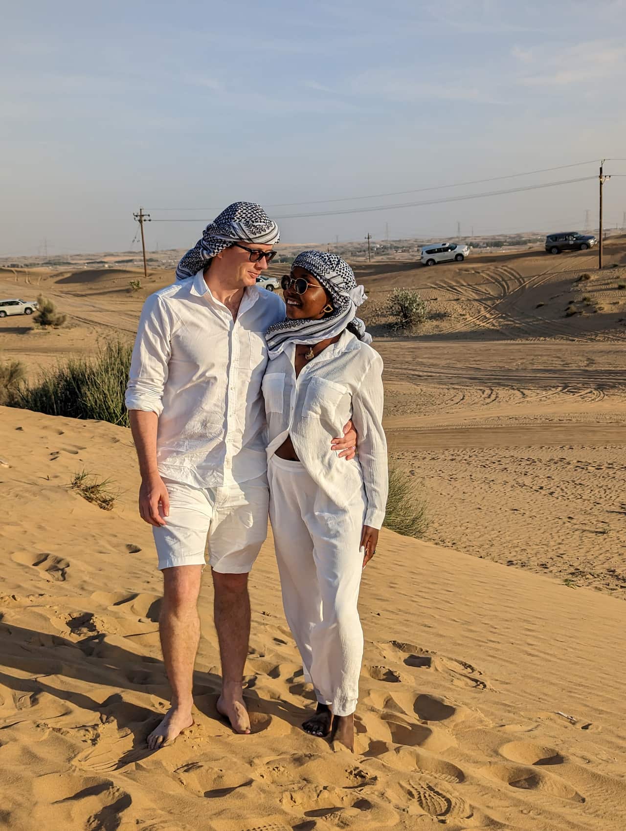 Man and woman dressed in white clothing stand on desert sand.