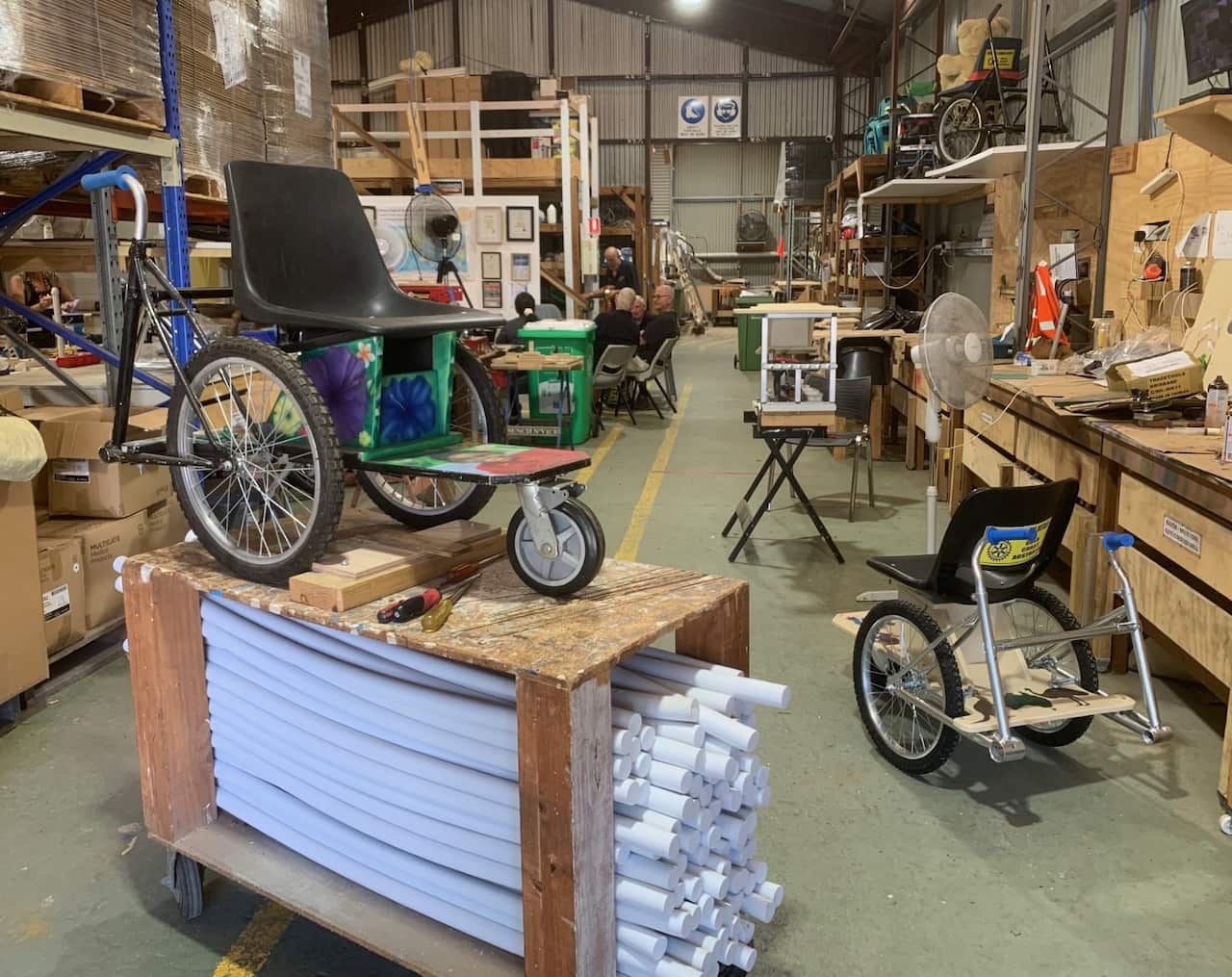 A wheelchair sits on a riser in a workshop with others in the background.