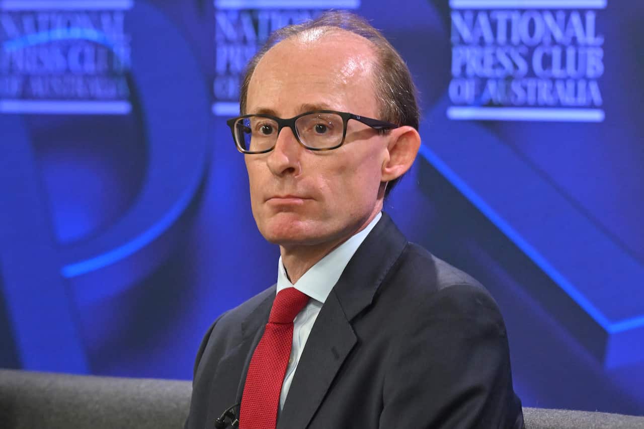 A man wearing glasses, a dark suit, and a red tie sits in front of a blue backdrop featuring the National Press Club of Australia logo.