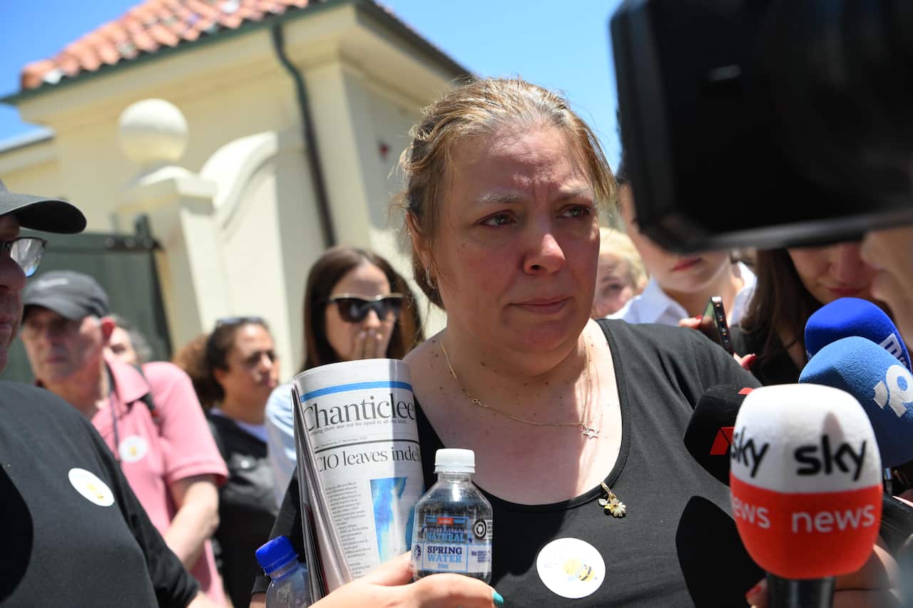 A woman standing outside, holding a newspaper, speaking into microphones.