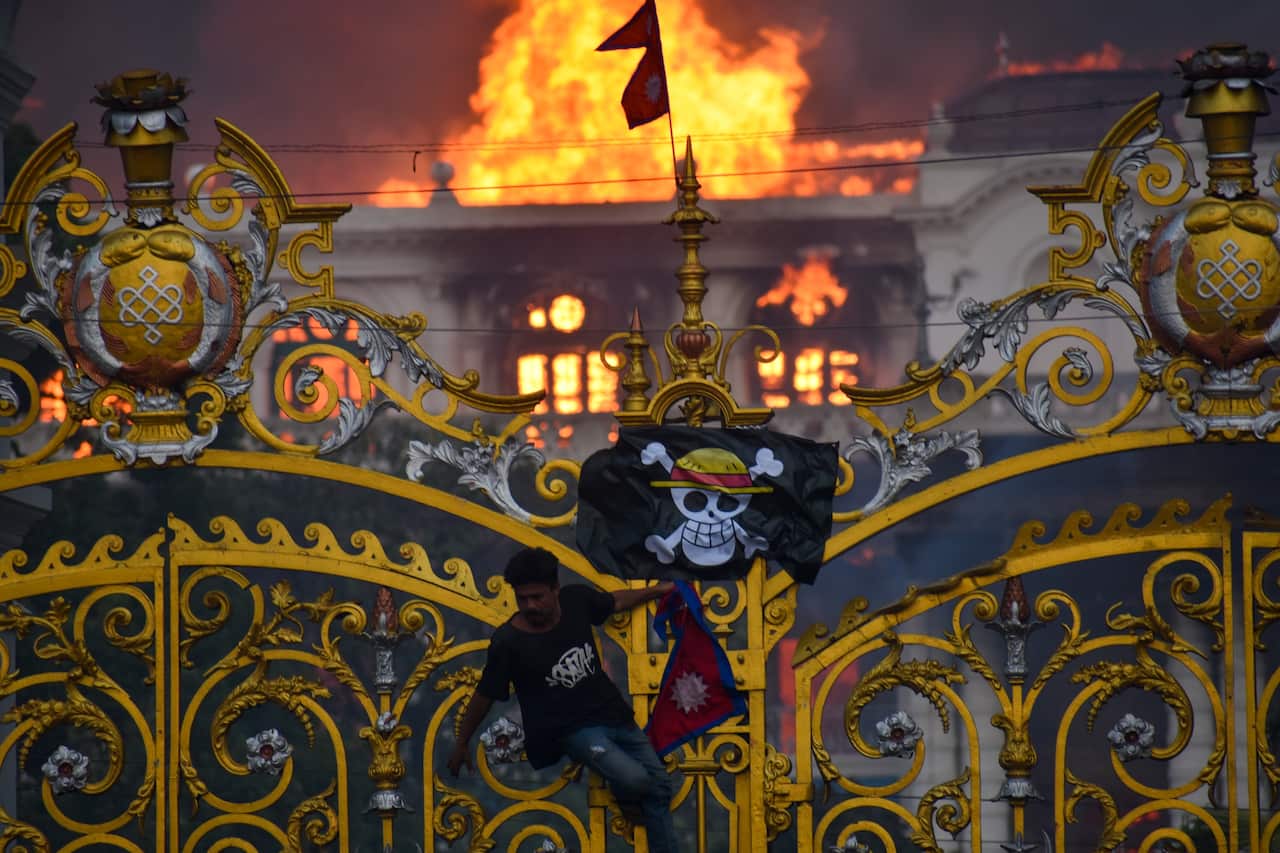 A large building is engulfed in flames behind a golden gate, with a man climbing the gate's bars. He is holding a Nepali flag and a pirate flag featuring a skull with a straw hat is hanging from the gate.