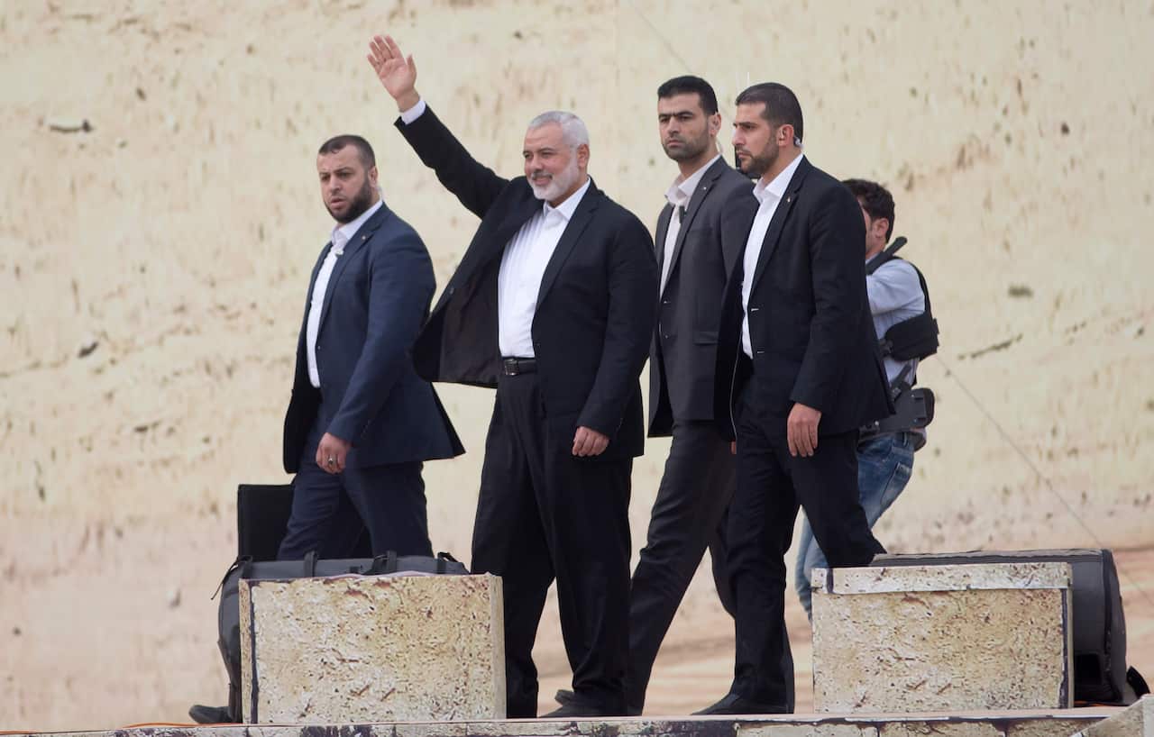 A group of men wearing suits walk together. One middle-aged man with a white beard raises his right hand.