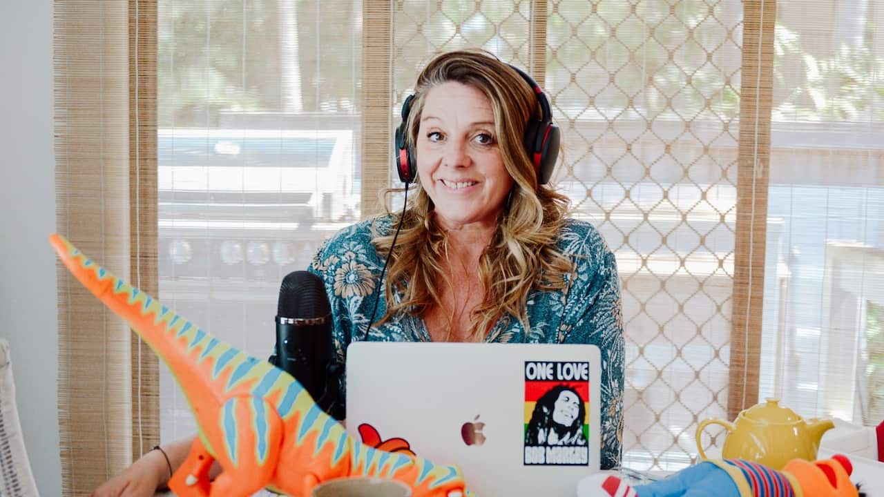 a woman with long blonde hair in a blue patterned dress sits at a table piled with toys and a laptop, podcast microphone and headphones