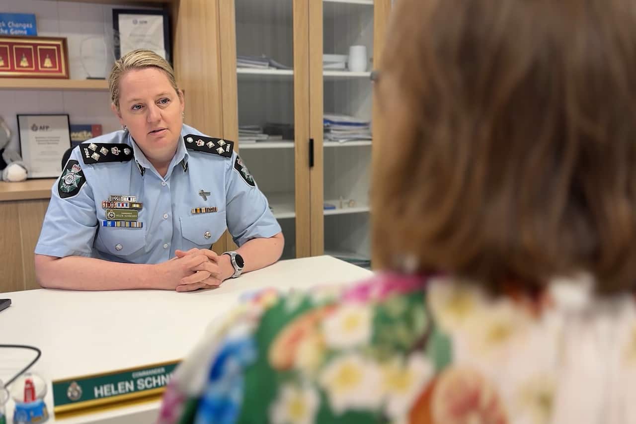 A female police officer in uniform speaks to another woman sitting across from her at a desk. 
