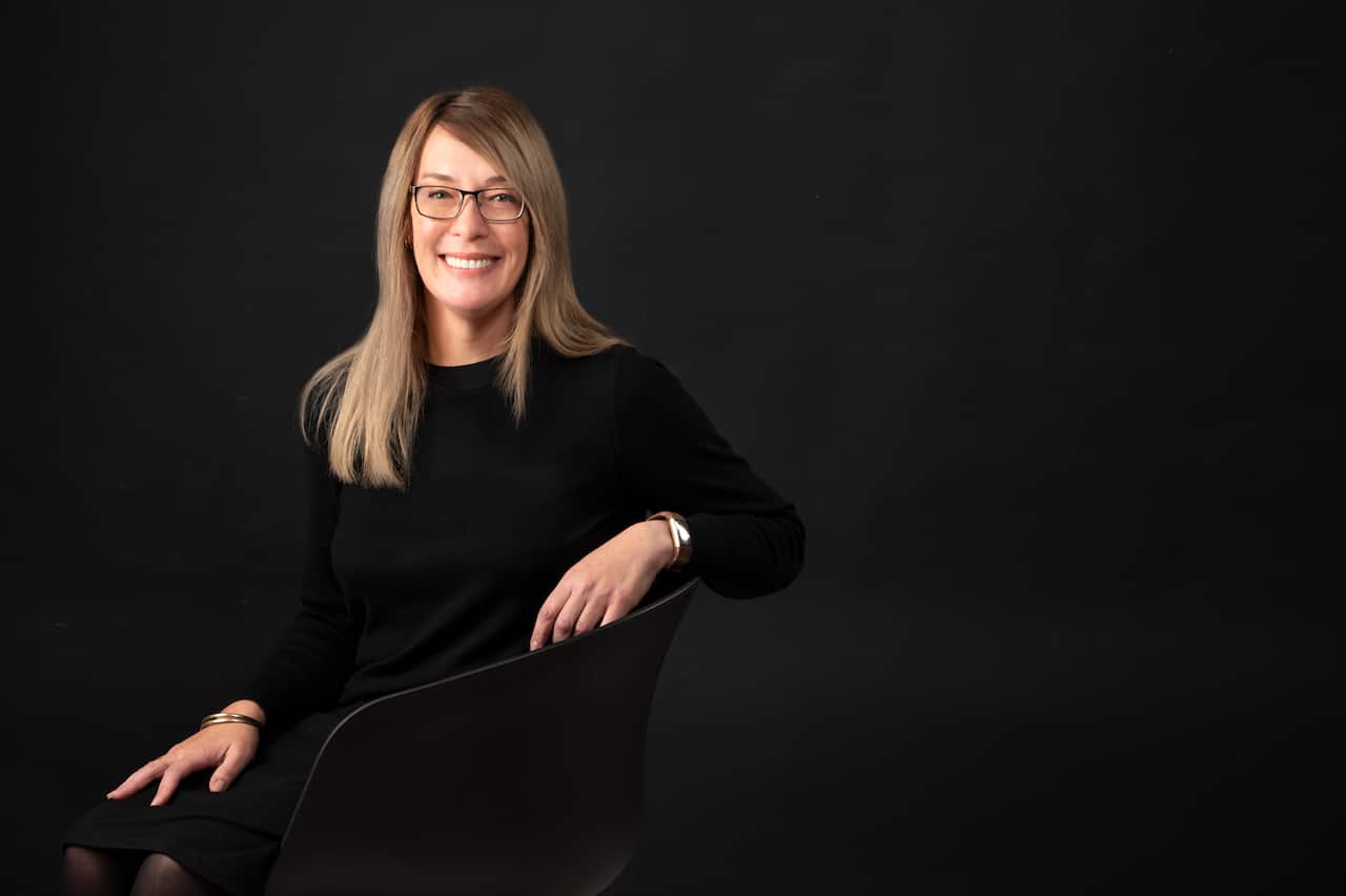 A woman dressed in black, wearing glasses, sits on a chair smiling.