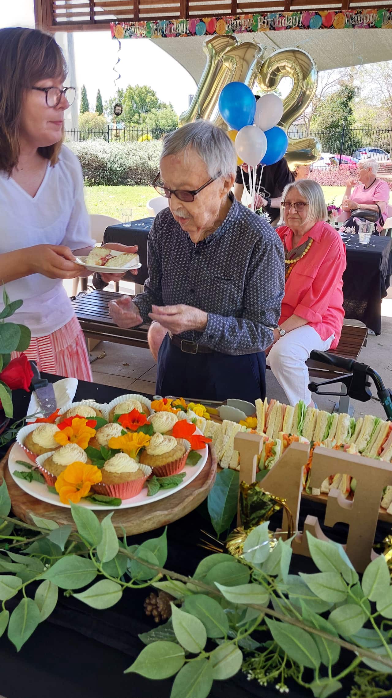 Ken Weeks, Australia's oldest man, stands by a table with sandwiches and cupcakes in front of gold balloons that say "112."
