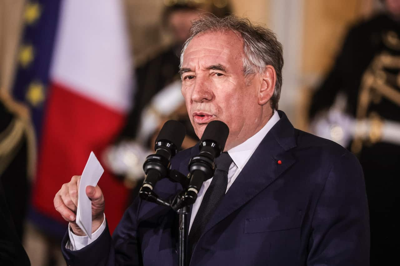 A man in a dark blue suit speaking into a microphone, holding a piece of paper in his hand.