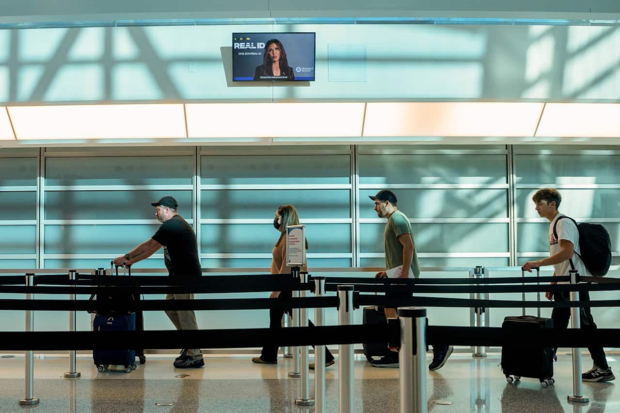 Four people wheel suitcases through the airport, above them is a screen displaying United States Secretary of Homeland Security Kristi Noem.
