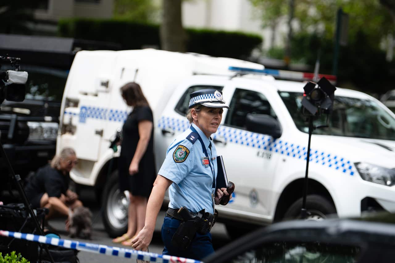 A police officer standing in front of a white and blue police car and a row of police tape.