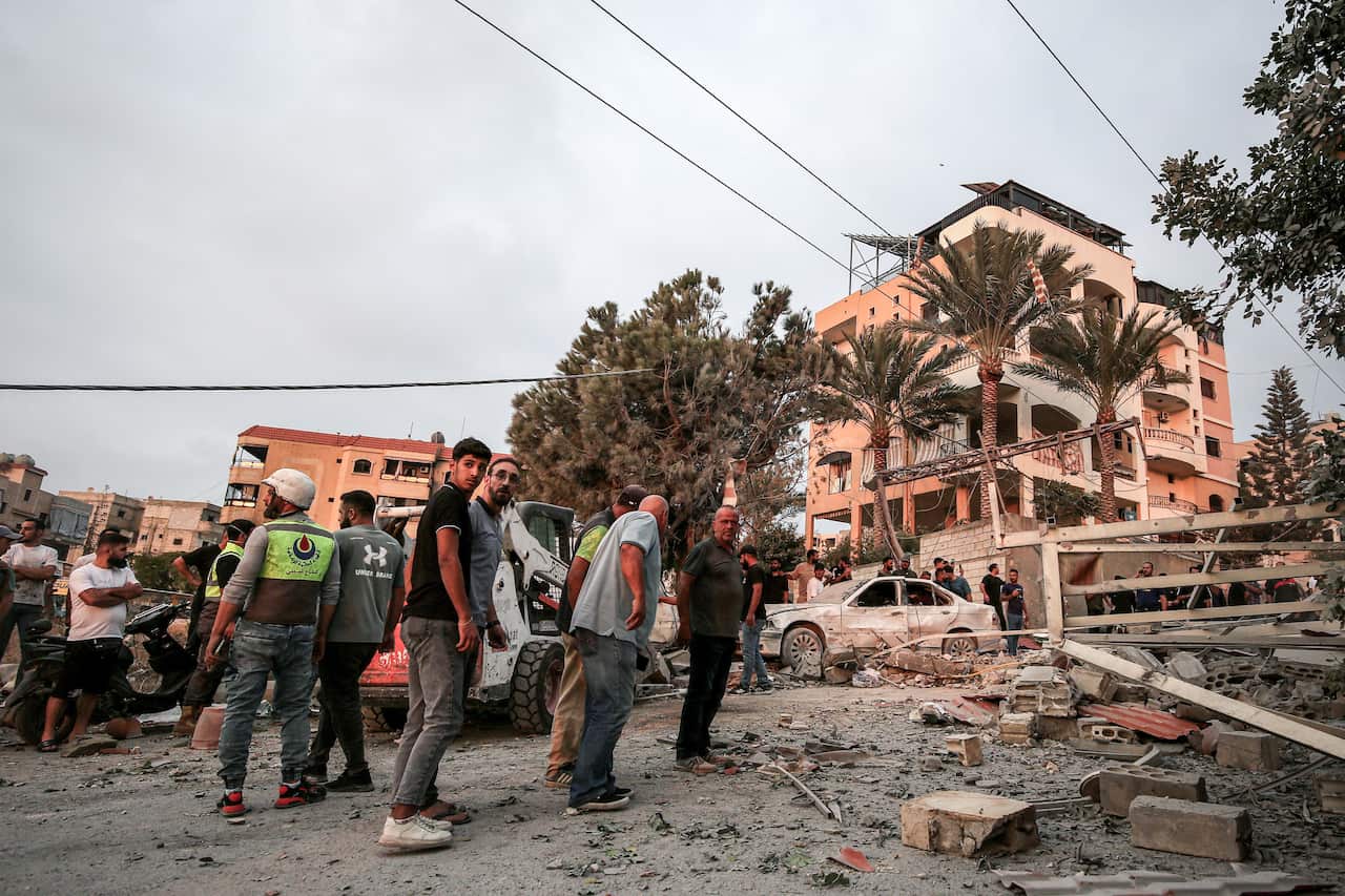 People survey destruction and rubble in an urban area, with a damaged white car and crumpled buildings in the background under an overcast sky.