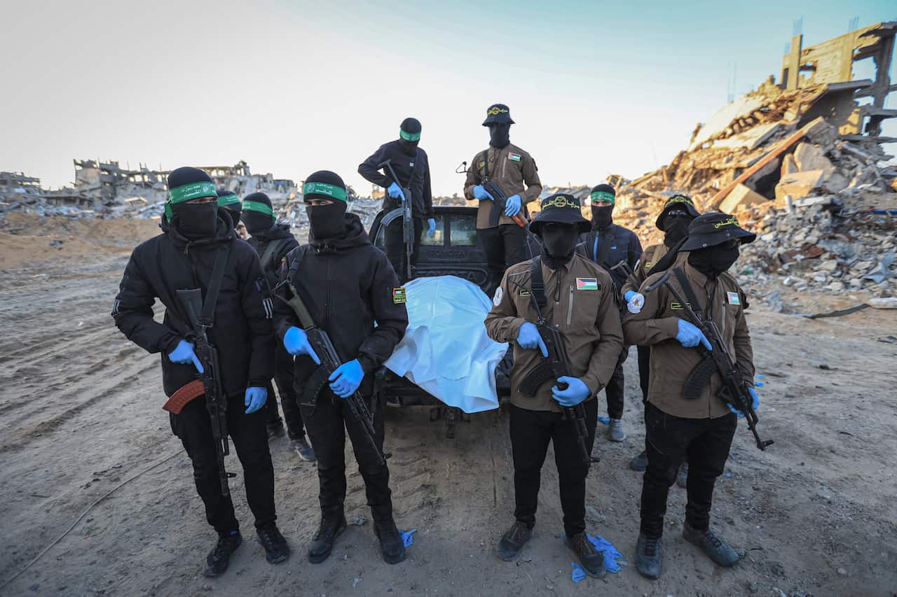Masked and armed militants in black and tan uniforms stand guard around a body wrapped in a white shroud on the back of a truck, set against a backdrop of heavily damaged buildings and rubble.