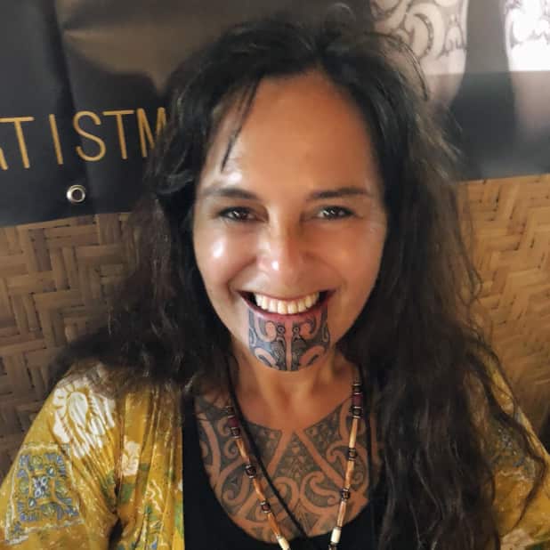 A woman with a facial tattoo smiling for a photo. She's wearing a yellow top and her chest tattoo is also visible.