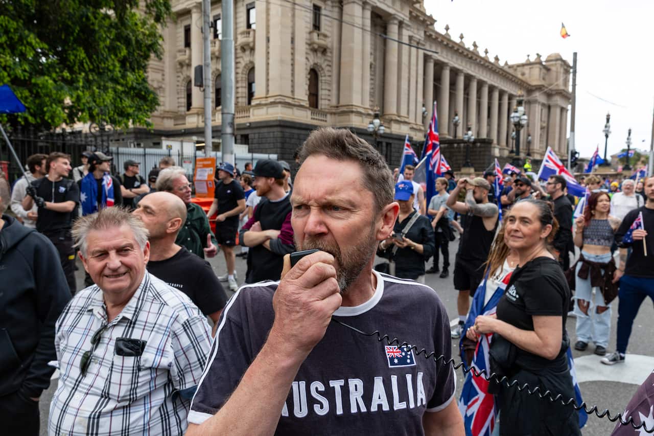 A man shouting into a handheld speaker during a protest march. There's a crowd behind him, many of whom are holding Australian flags.