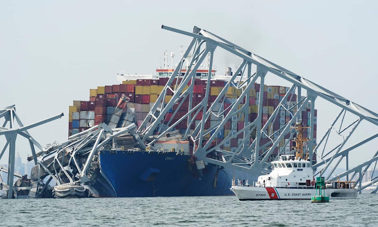 A small white Coast Guard cutter passes in front of a container ship with collapsed sections of a metal bridge laying over the bow.