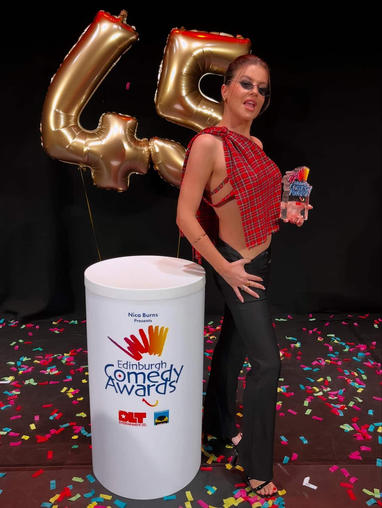 A woman wearing a red tartan top and black trousers poses with an award trophy. Behind her are gold balloons in the shape of the number 45
