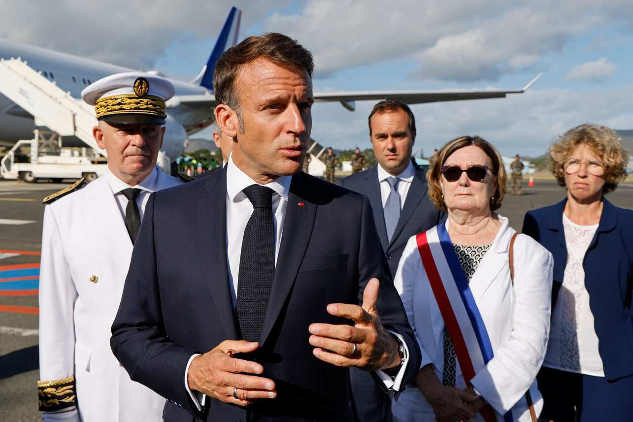French President Emmanuel Macron gestures with left hand as members of his delegation stand behind him.
