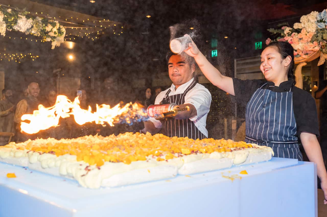 Um chef usa um maçarico para caramelizar uma sobremesa grande e outro polvilha com pó.