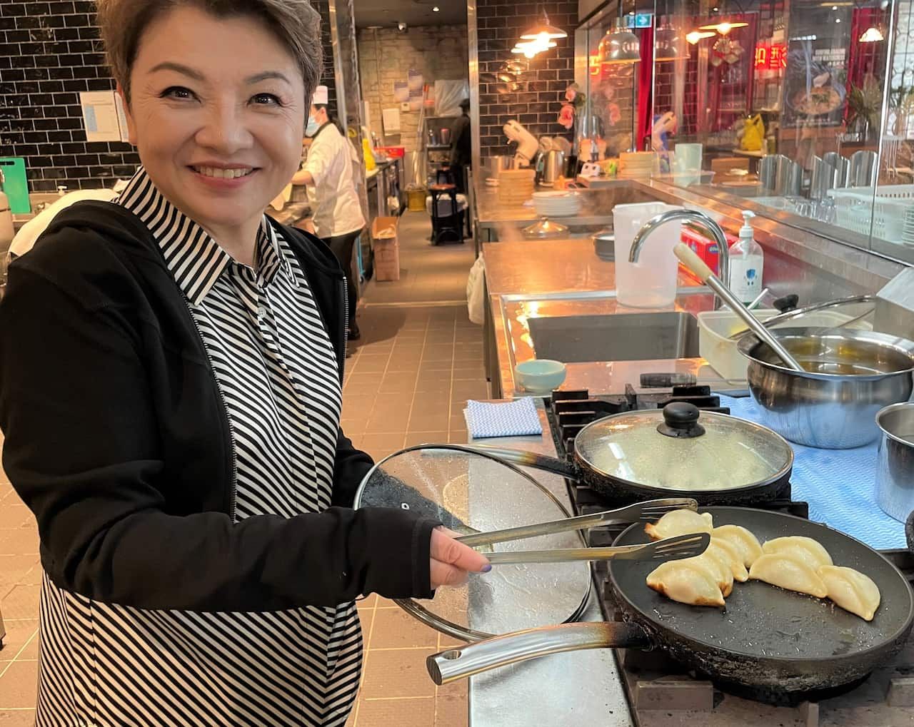 A woman in a striped shirt and black jacket stands over a hot plate cooking dumplings. 