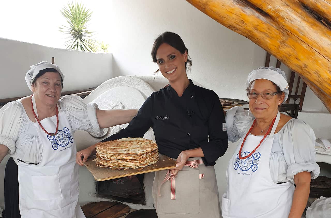 A young woman in a black chef's jacket stands holding a tray of flatbread. She is flanked by two women in white shirts and aprons.