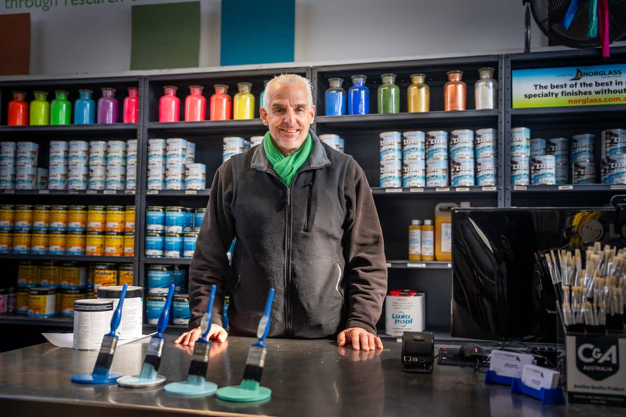 A man in a grey sweater stands at a counter in front of shelves of paint.