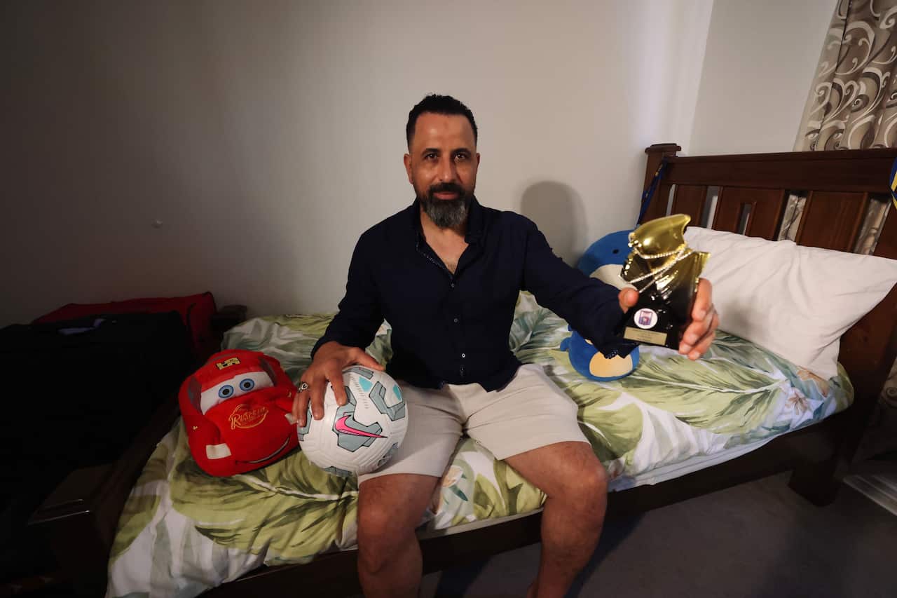 A man sitting on a children's bed, holding a soccer ball and trophy.