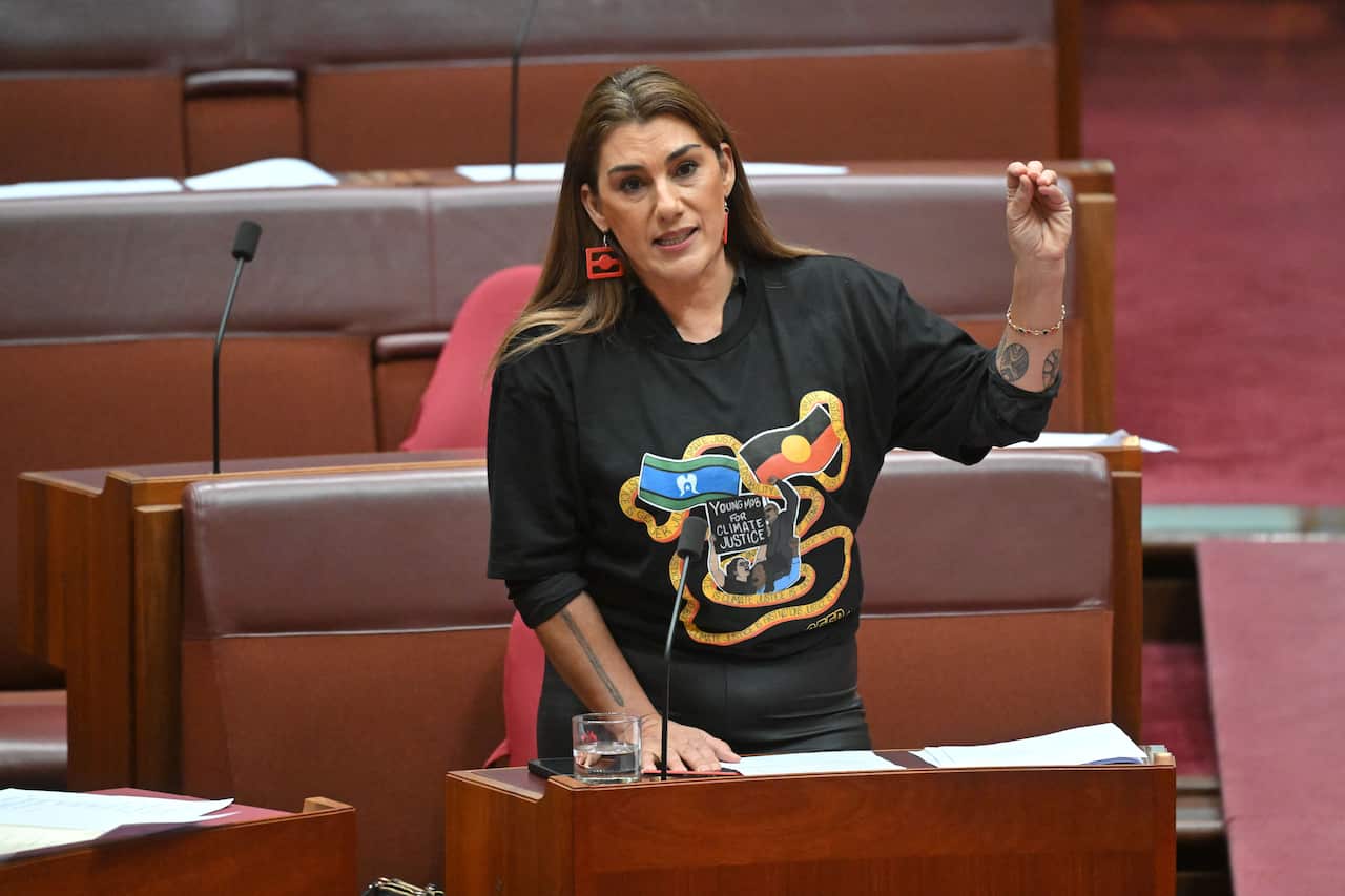 Independent senator Lidia Thorpe in parliament, speaking with her hand raised and wearing a black T-shirt depicting a logo promoting climate justice that includes the Aboriginal and Torres Strait Islander flags.