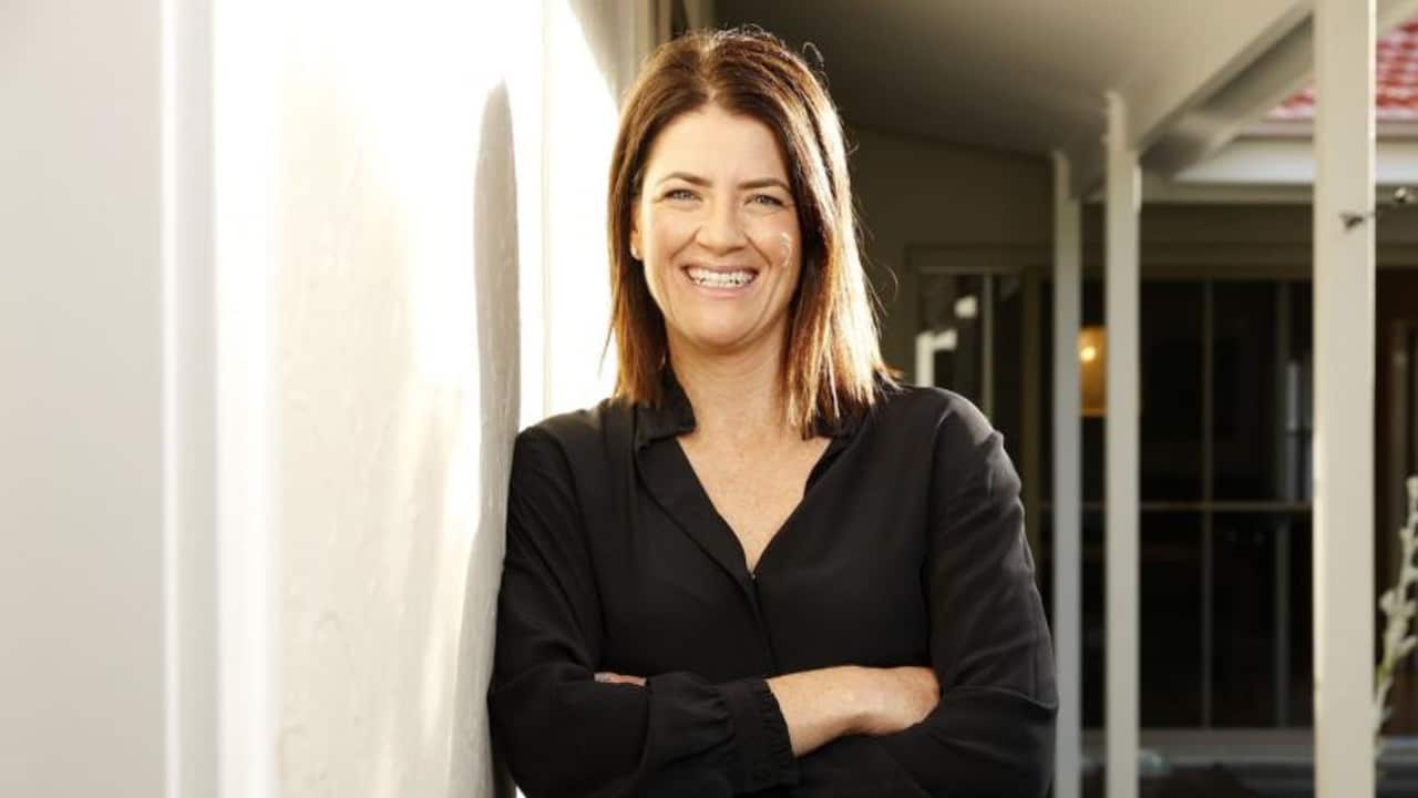 a woman with light brown shoulder length hair and in a black top stands leaning outside on a white wall and smiles at the camera