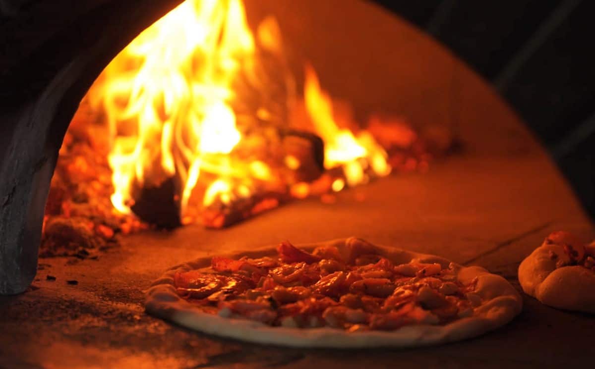 A pizza topped with salami sits on a stone slab in a wood-fired oven.