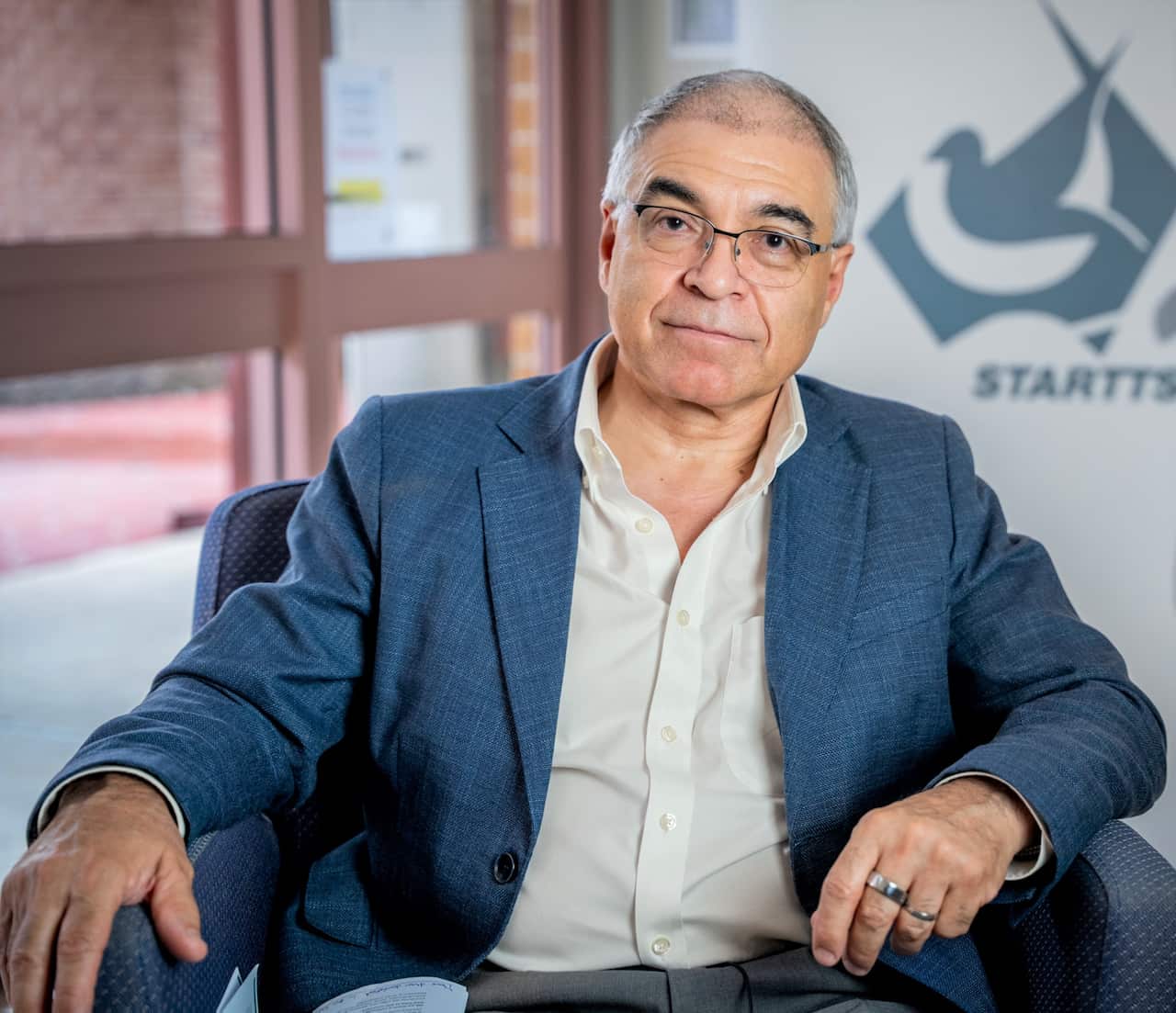 A man wearing a blue jacket sits on a chair in front of a sign saying STARTTS.