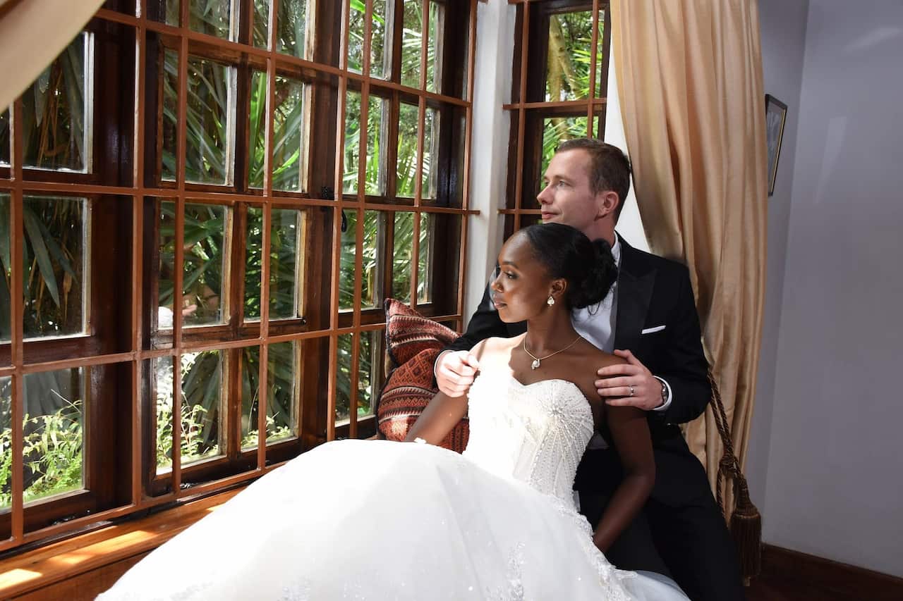 A woman in a white dress sits in front of a man in a suit who is holding her shoulders. The two are looking out a window.