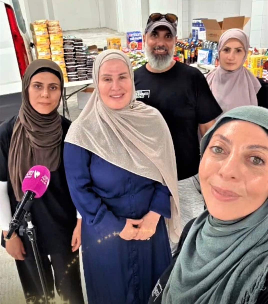 A woman in a blue dress stands at the centre of a group, with food products on a shelf bhind them. 