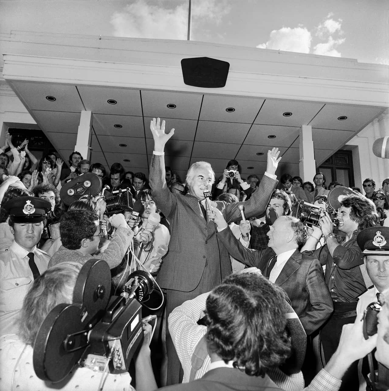 A black and white photograph of Gough Whitlam on the steps of old parliament, with both hands up in the air and a microphone to his face, surrounded by reporters.