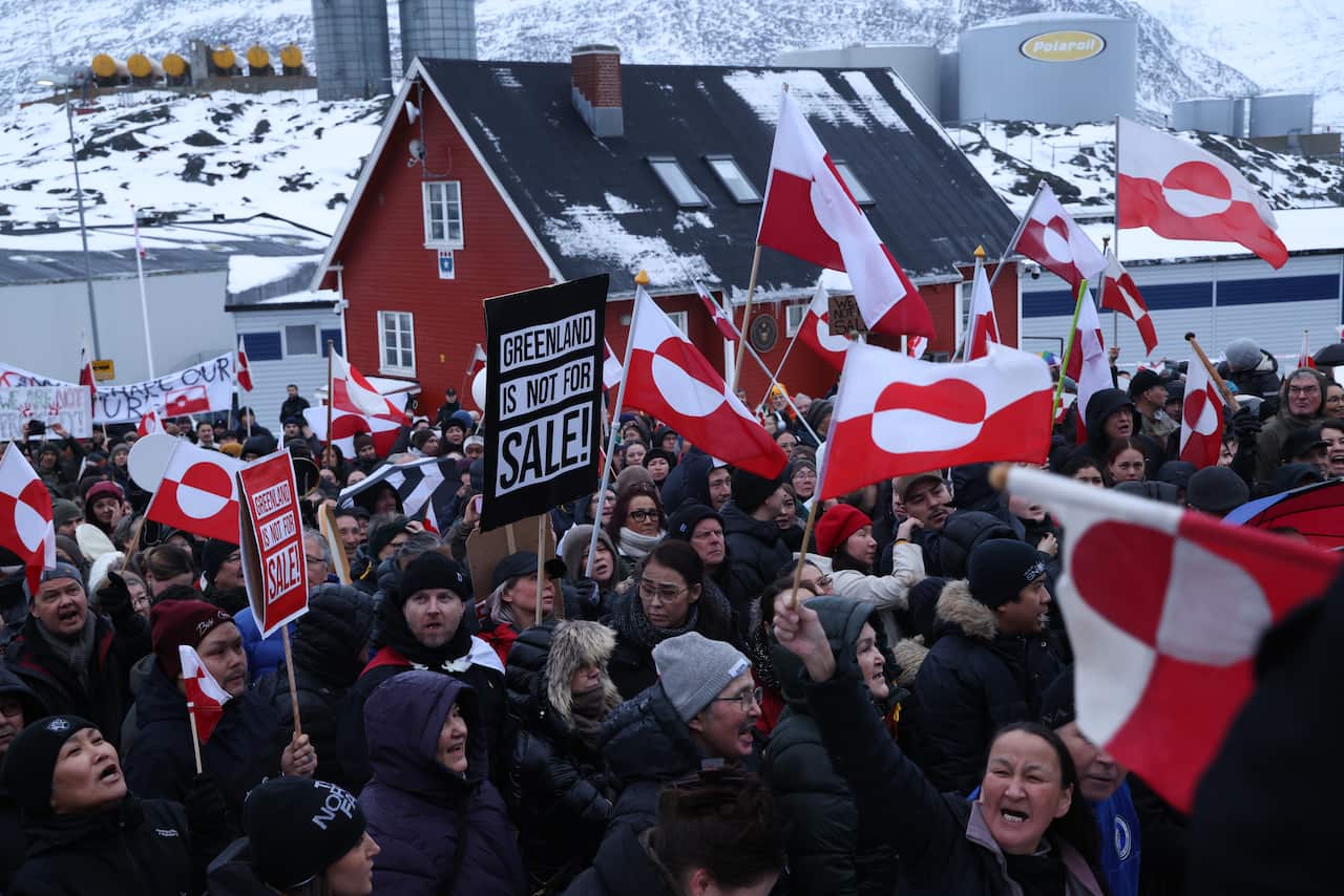 Protesters carry red and white flags and signs saying "Greenland is not for sale" in a snowy area. 
