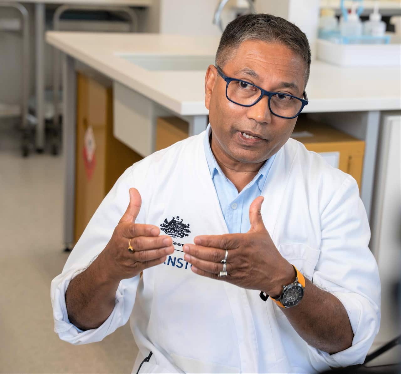 A man with brown hair wearing a white lab coat sits in a science lab holding his hands up.