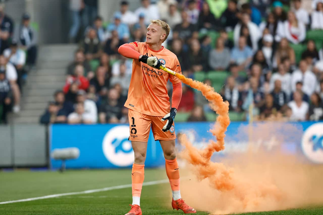 A goalkeeper dressed in orange throws a flare with orange smoke coming from it.