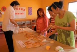 A man in a white shirt is standing in front of a low, long table laden with bread loaves, with three women standing on the opposite side touching the loaves.