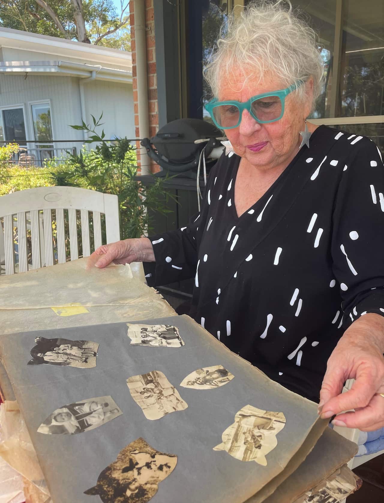 A woman in green glasses sits at a table looking at an album of old photos.