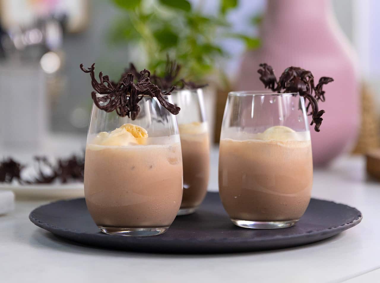 Three glasses sit on a round dark tray. The glasses are three-quarters filled with a chocolate drink, and each glass is garnished with a chocolate twirl.