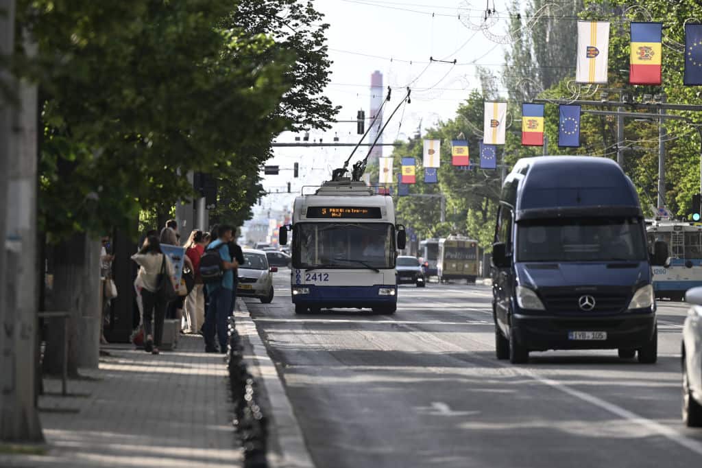 A trolley bus and van drive along a busy road. Pedestrians walk on a footpath beside the road.
