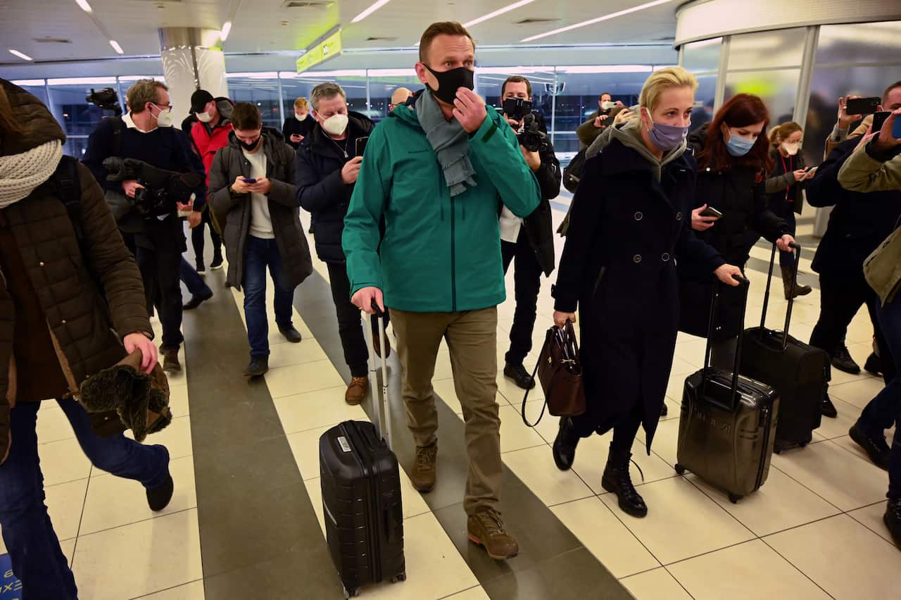 A group of people with luggage, led by a man in a green jacket and a face mask, are walking through the airport.