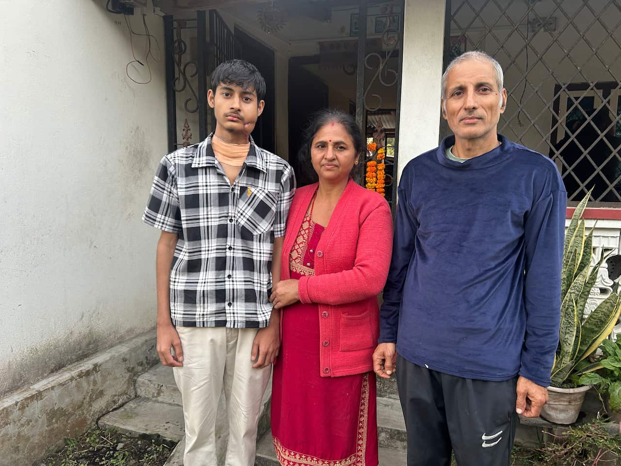 A young man with a large bandage around his neck and a scar on the left side of his lower jaw stands outside a house. His mother, who is dressed in red, has her hand on his arm. Next to her is his father. They all have neutral facial expressions. 