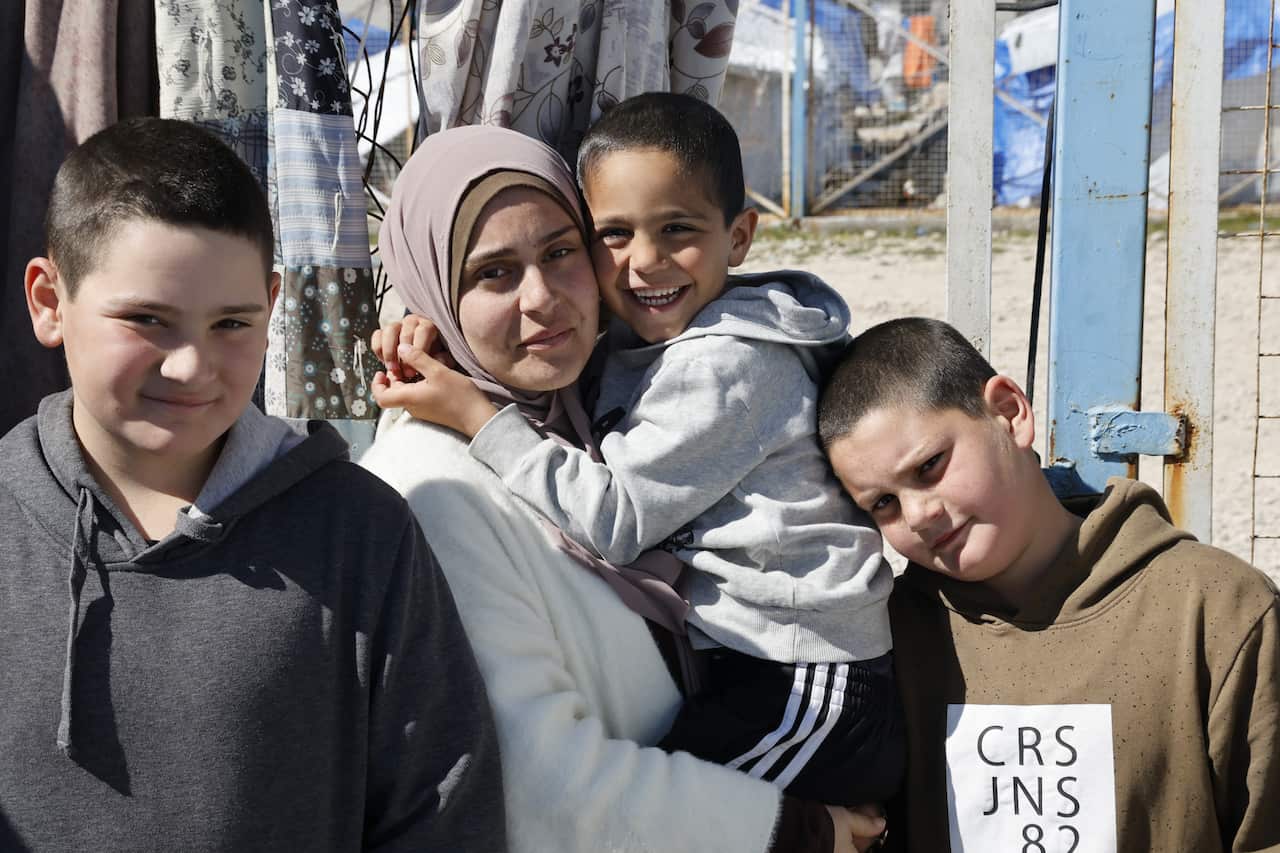 A woman in a hijab holds a young boy, who is grinning. To her left and right are two older boys. One has a slight smile, and one has a neutral facial expression. 