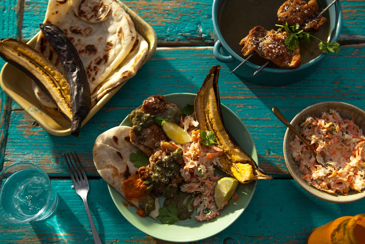 A pale green bowl with curry, a piece of plantain, a flatbread and salad sits on a blue wooden table. Other bowls holding more bread, salad and curry sit nearby on the table.