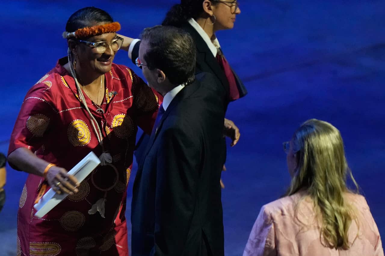 An Indigenous woman wearing a red patterned dress and a feathered headband smiles warmly while greeting a man in a dark suit at a formal event.