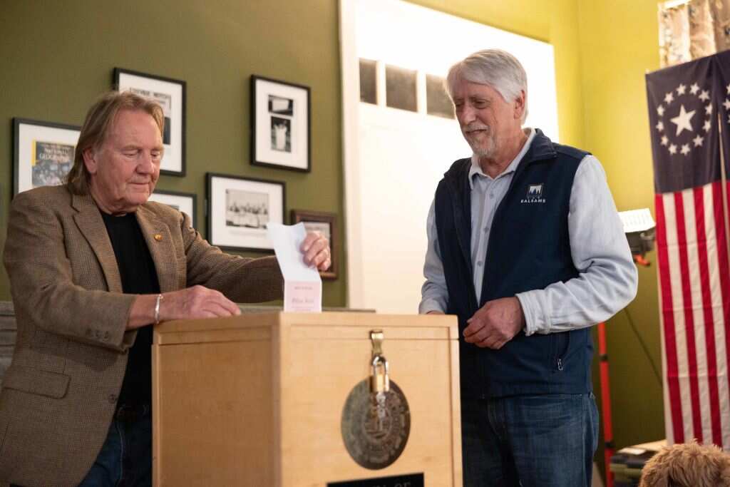 A man puts a piece of paper in a wooden box with a lock on it. Another man is watching him.