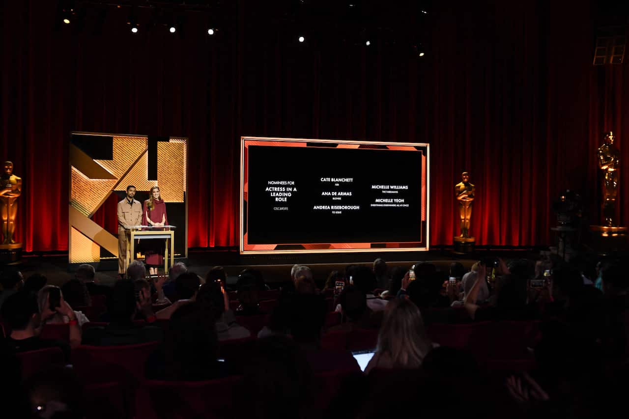 Two people in the theater on the stage behind the pulpit. The big screen next to them displays the names of people. The theater has seated audiences.