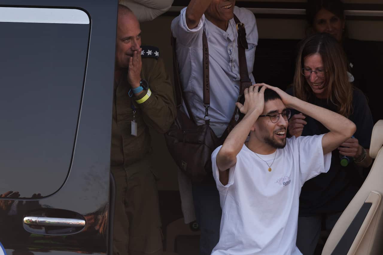 A young man in a white t-shirt displays an expression of shock and joy with his hands on his head as he is greeted by smiling individuals and a uniformed official near a vehicle.