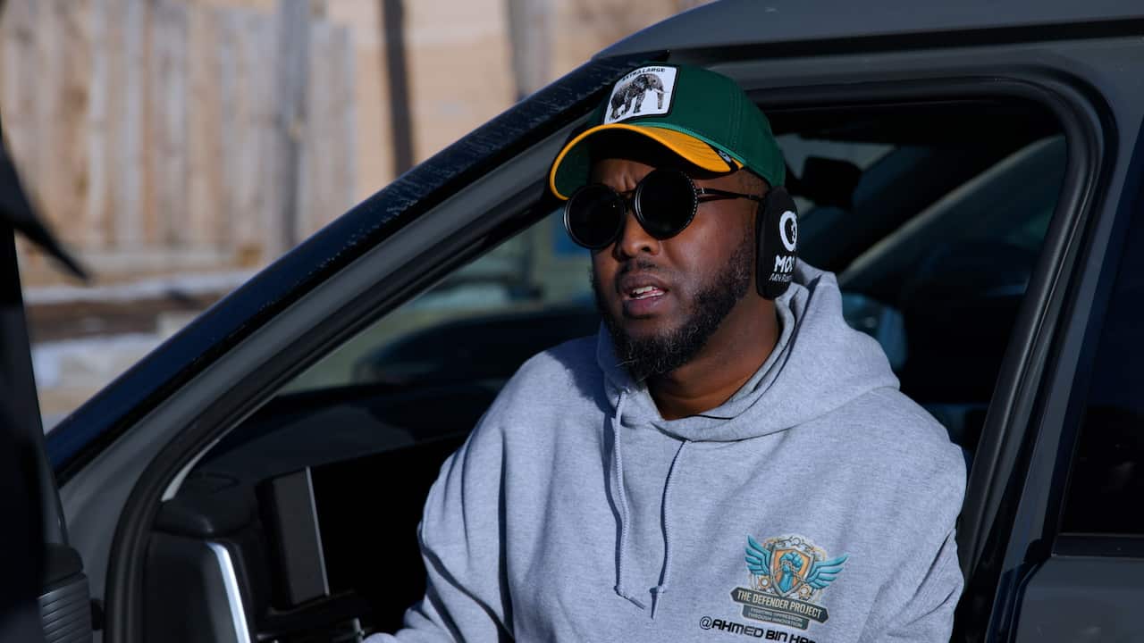 A man wearing sunglasses, a baseball cap and earmuffs sits in a car with the door open.